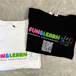Haft z radości - nadruki i haft na odzieży Warszawa - Dwie złożone koszulki: biała i czarna z kolorowym nadrukiem 'FUN&LEARN Akademia Kreatywności', na czarnej koszulce dodatkowo kod QR.