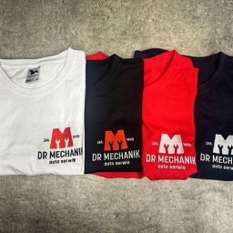 Haft z radości - nadruki i haft na odzieży Warszawa - Cztery kolorowe koszulki z krótkim rękawem (biała, czarna, czerwona, granatowa) leżące na szarym tle, każda z nadrukowanym logo warsztatu samochodowego 'Dr Mechanik'.