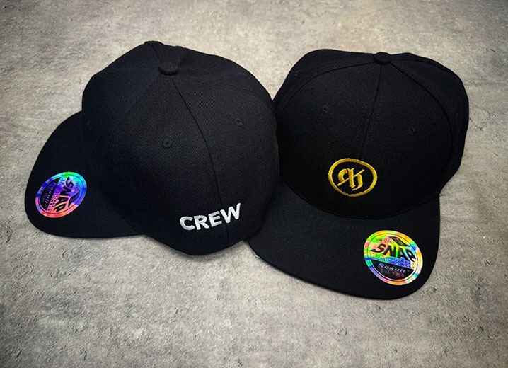 Dwie czarne czapki typu snapback leżące na szarym tle, jedna z napisem 'CREW' z tyłu, druga z wyhaftowanym złotym logo na przodzie i holograficzną naklejką na daszku.