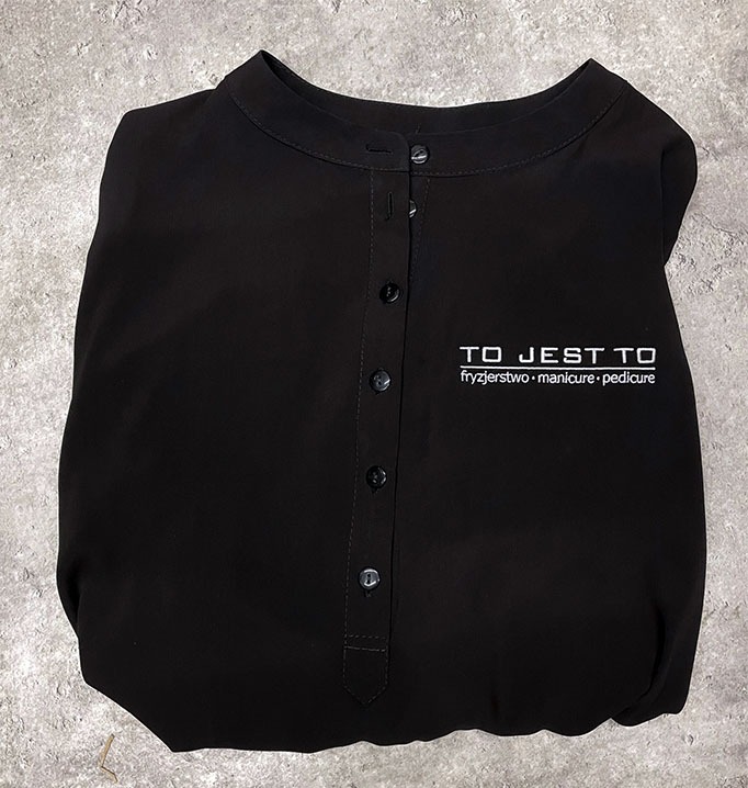 Czarna koszula z długim rękawem, zapinana na guziki, z wyhaftowanym logo 'TO JEST TO fryzjerstwo • manicure • pedicure' na piersi, leżąca na szarym tle.
