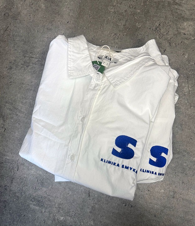 Biała koszula z długim rękawem, złożona na szarym tle, z niebieskim nadrukiem logo 'S Klinika Smyka'