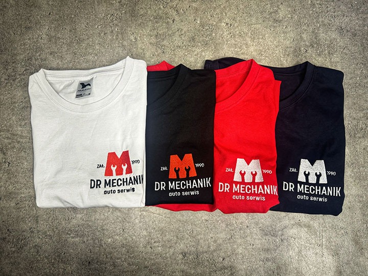 Cztery kolorowe koszulki z krótkim rękawem (biała, czarna, czerwona, granatowa) leżące na szarym tle, każda z nadrukowanym logo warsztatu samochodowego 'Dr Mechanik'.