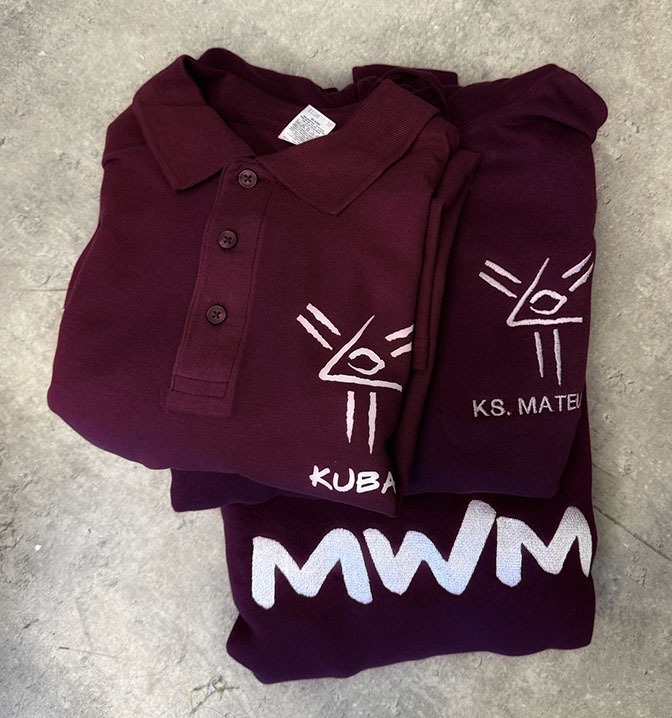 Stos bordowych koszulek polo z białymi nadrukami, w tym logo z okiem w trójkącie i napisami 'KUBA', 'KS. MATEU' i 'MWM', leżących na betonowej posadzce.