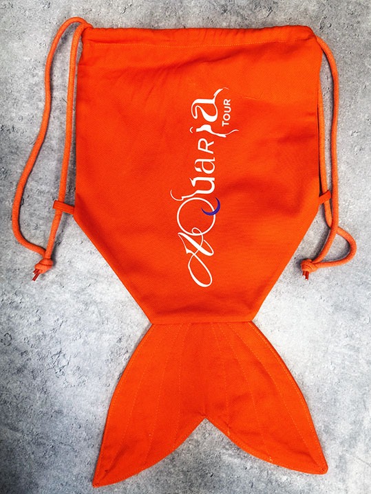 Pomarańczowy worek ze sznurkiem w kształcie syreniego ogona z białym nadrukiem 'Aquaria TOUR' na szarym tle.