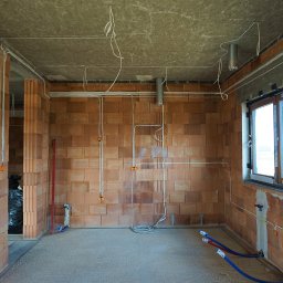 PR-System - Surowy stan wnętrza z widoczną instalacją elektryczną w peszlach i puszkach, częściowo zabetonowaną podłogą, oraz rurami wodnymi przy ścianie z oknem.