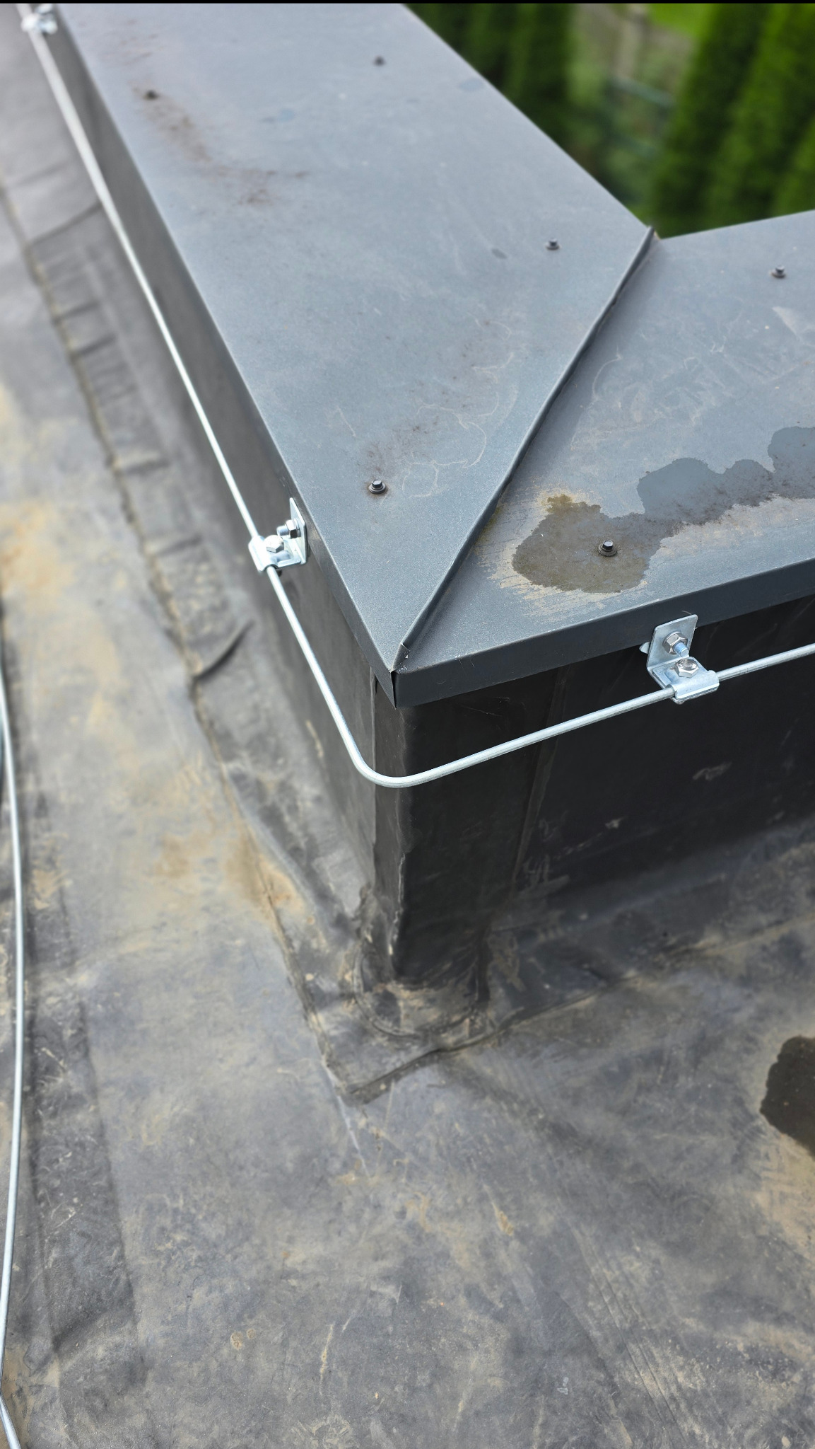 Metalowa obudowa komina na dachu pokrytym papą, zabezpieczona odgromowo za pomocą białego przewodu i metalowych uchwytów.