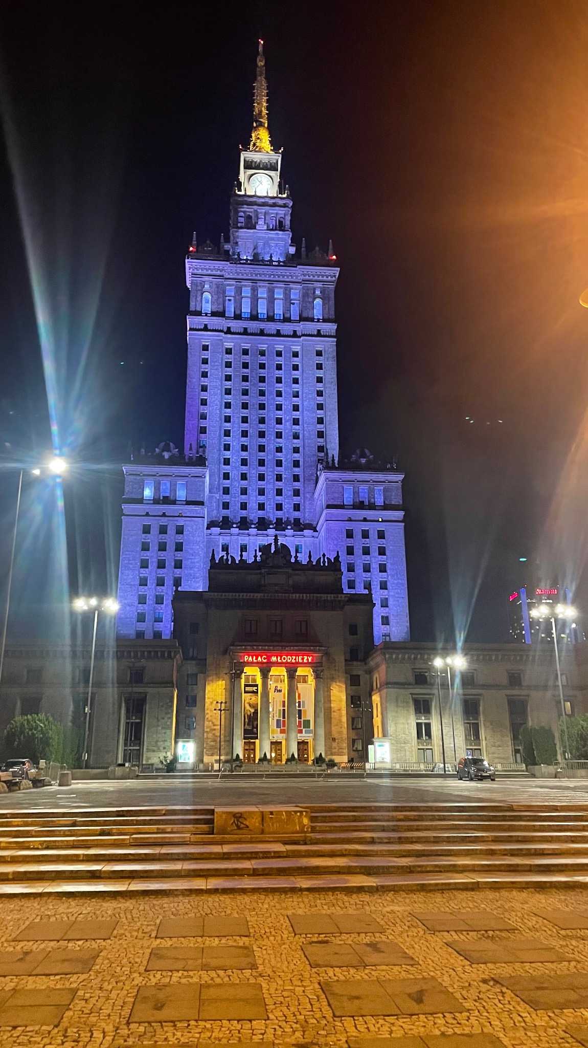 Pałac Kultury i Nauki w Warszawie oświetlony na niebiesko w nocy, widok od strony schodów z kostki brukowej.