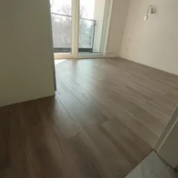 Ułożone panele podłogowe w jasnym, pustym pokoju z balkonem, widoczne ślady po niedawno zakończonych pracach wykończeniowych.