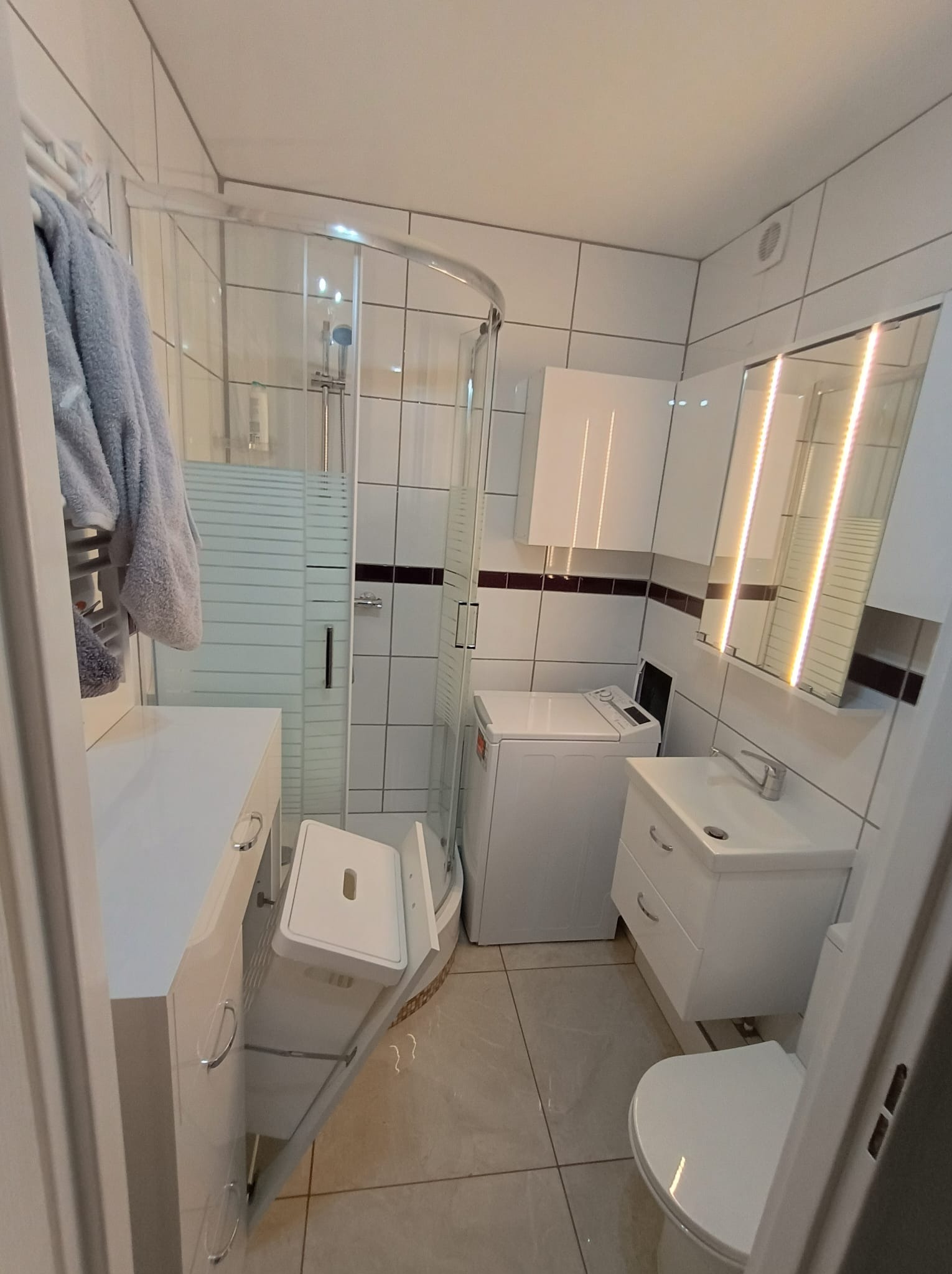 Jasna łazienka z kabiną prysznicową narożną, pralką, umywalką z szafką i toaletą. Otwarty kosz na pranie w szafce pod umywalką. Lustro z oświetleniem LED.