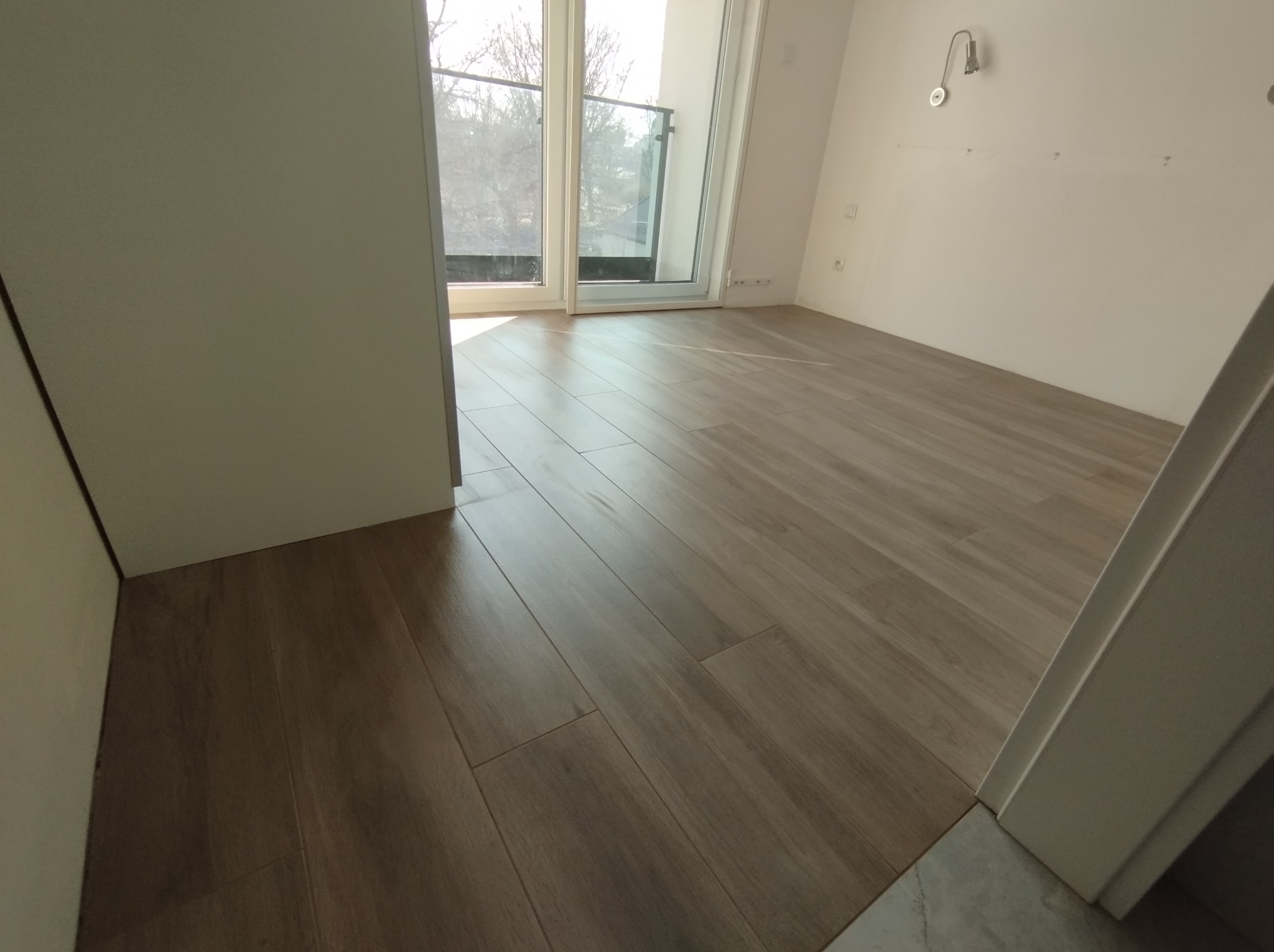 Ułożone panele podłogowe w jasnym, pustym pokoju z balkonem, widoczne ślady po niedawno zakończonych pracach wykończeniowych.