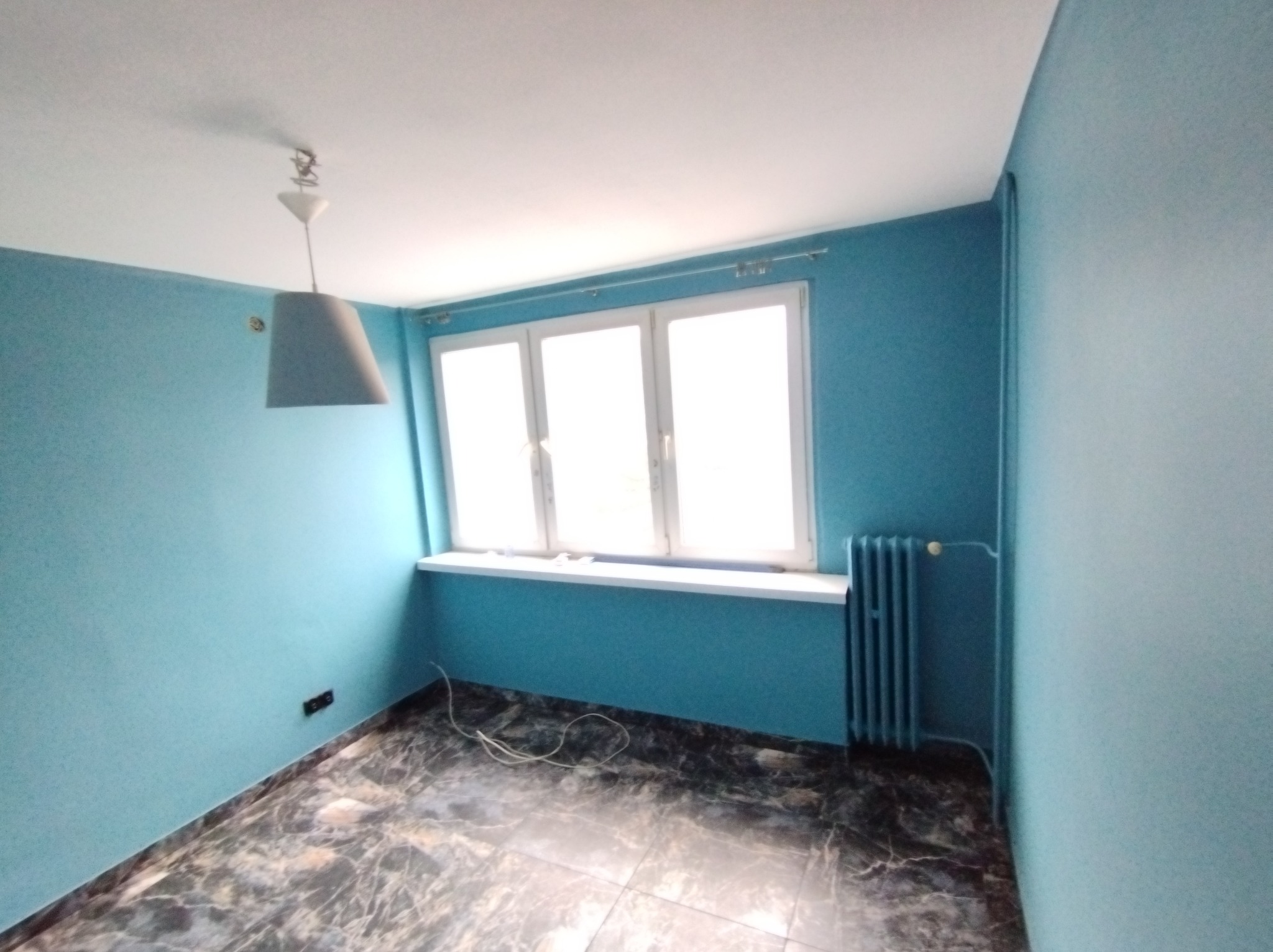 Pomalowane na turkusowo wnętrze pokoju z marmurkową podłogą, białą stolarką okienną i szarym kloszem lampy wiszącej.