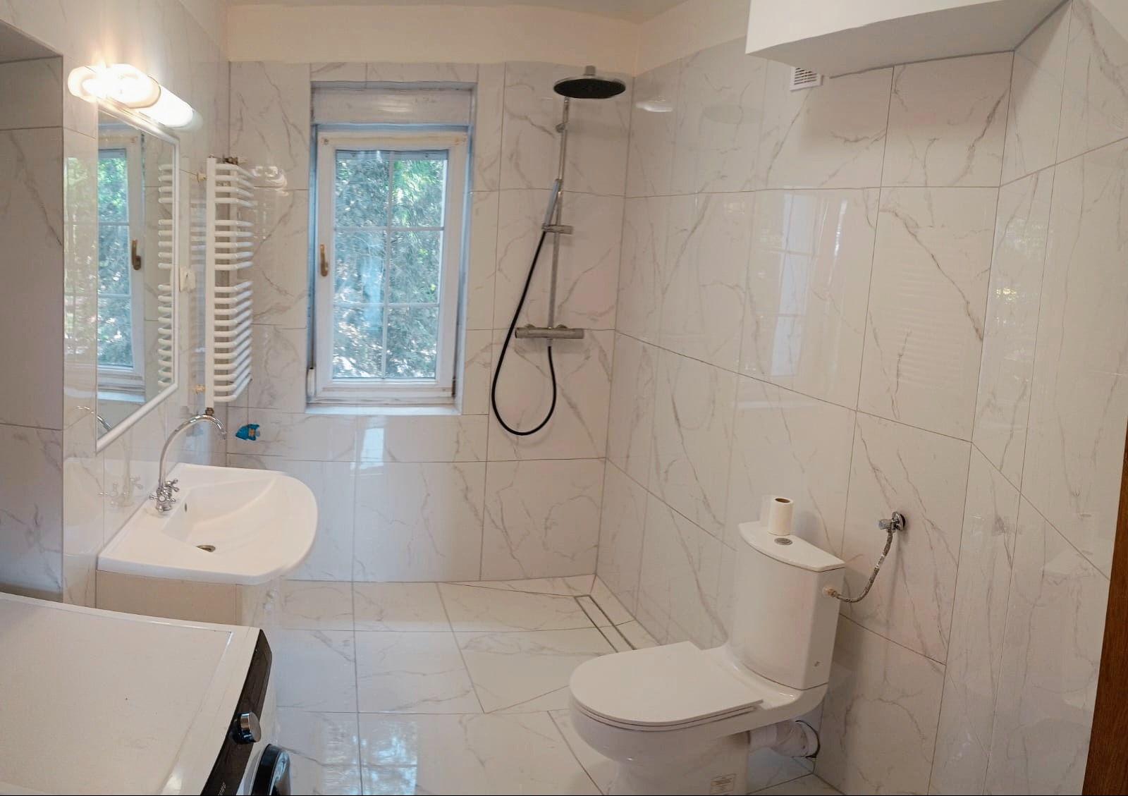 Jasna łazienka z marmurowymi płytkami, kabiną prysznicową, umywalką i grzejnikiem drabinkowym. Okno z widokiem na zieleń. Minimalistyczny design.