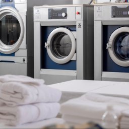 Abrilux - Wyposażenie Pralni - Rząd profesjonalnych pralek Electrolux w pralni przemysłowej, z stosem czystych, białych ręczników na pierwszym planie.