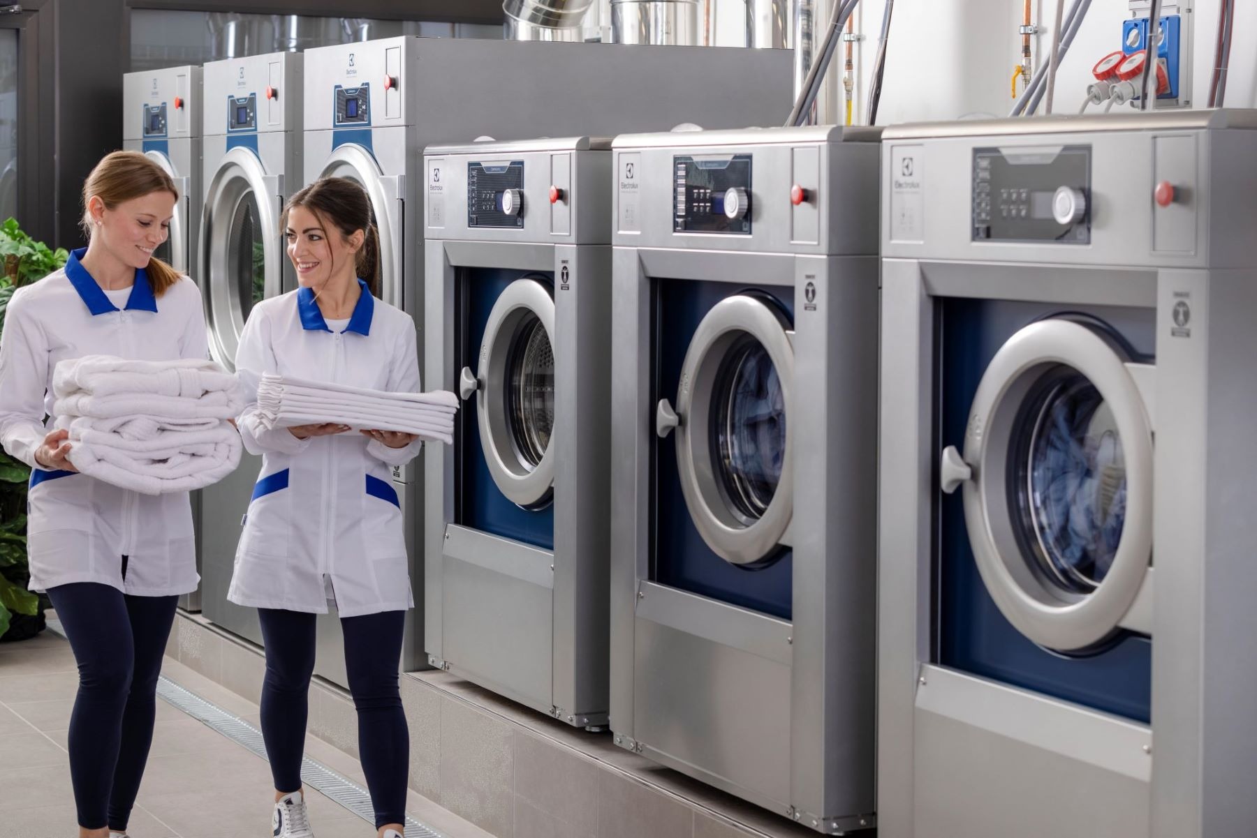 Dwie kobiety w białych uniformach z niebieskimi akcentami niosą sterty czystych, białych ręczników na tle rzędu profesjonalnych pralek Electrolux w pralni.