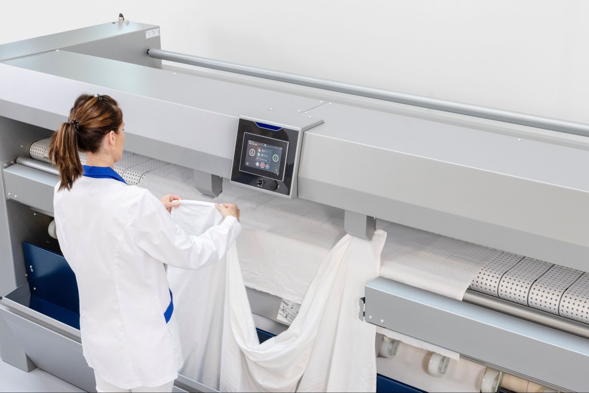 Pracownica w białym uniformie obsługuje przemysłową magielnicę, wkładając do niej białe prześcieradło.