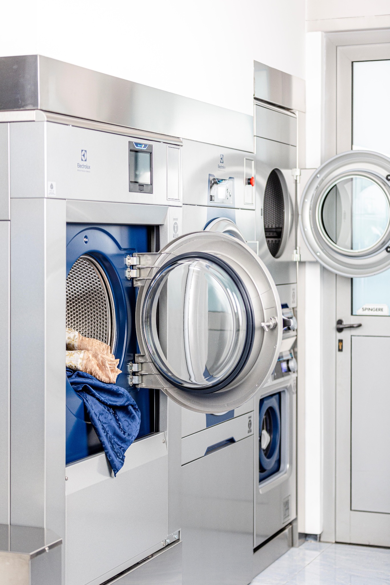 Rząd profesjonalnych pralek przemysłowych Electrolux z otwartymi drzwiami, bęben jednej z nich załadowany częściowo praniem, w tle widoczne drzwi i kolejne pralki