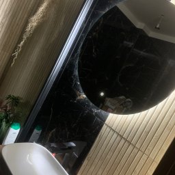 Wojtek gielarowski - Umywalka nablatowa REA w łazience z czarnym marmurem i drewnopodobnymi płytkami ściennymi, okrągłe podświetlane lustro odbija sufit.