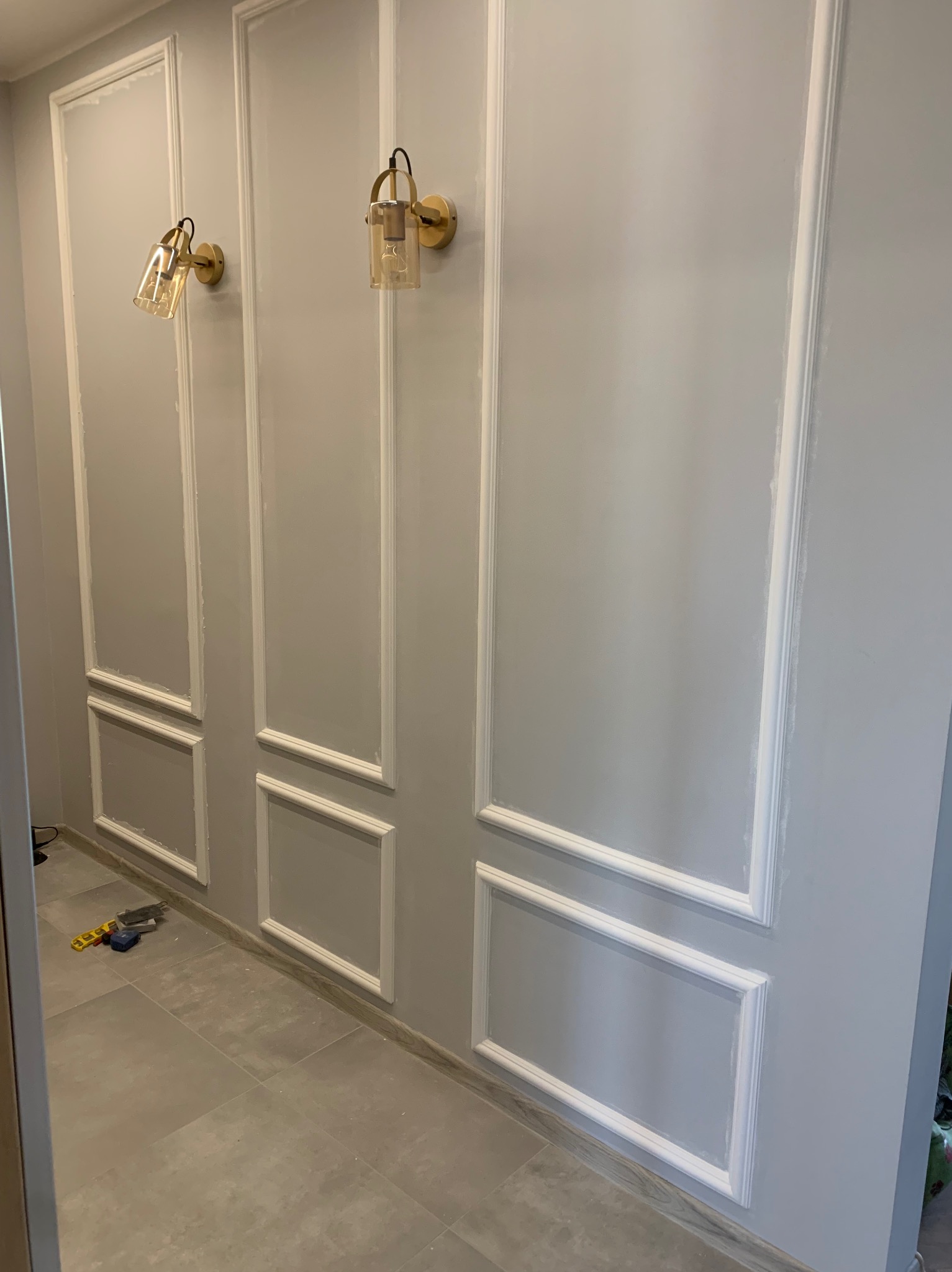 Szara ściana z dekoracyjnymi, białymi listwami w kształcie paneli i złotymi kinkietami. Na podłodze leżą narzędzia.