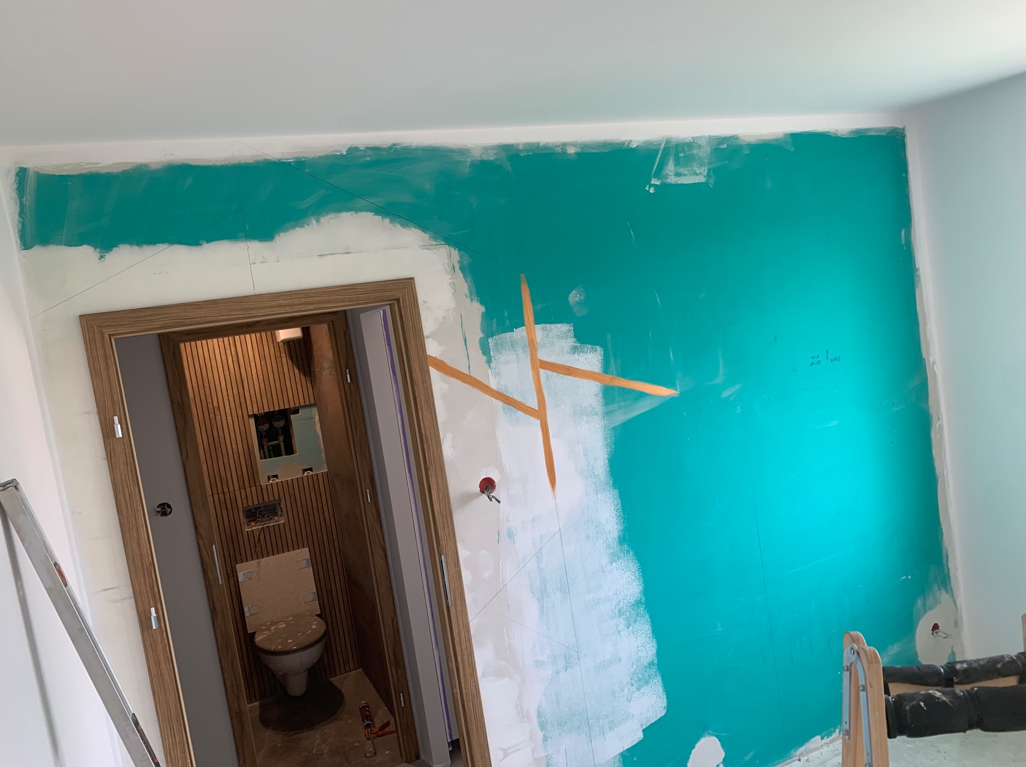 Ściana w trakcie malowania na turkusowo, widoczne ślady po taśmie malarskiej i niedomalowane fragmenty, obok wejście do łazienki z drewnianymi panelami ściennymi.