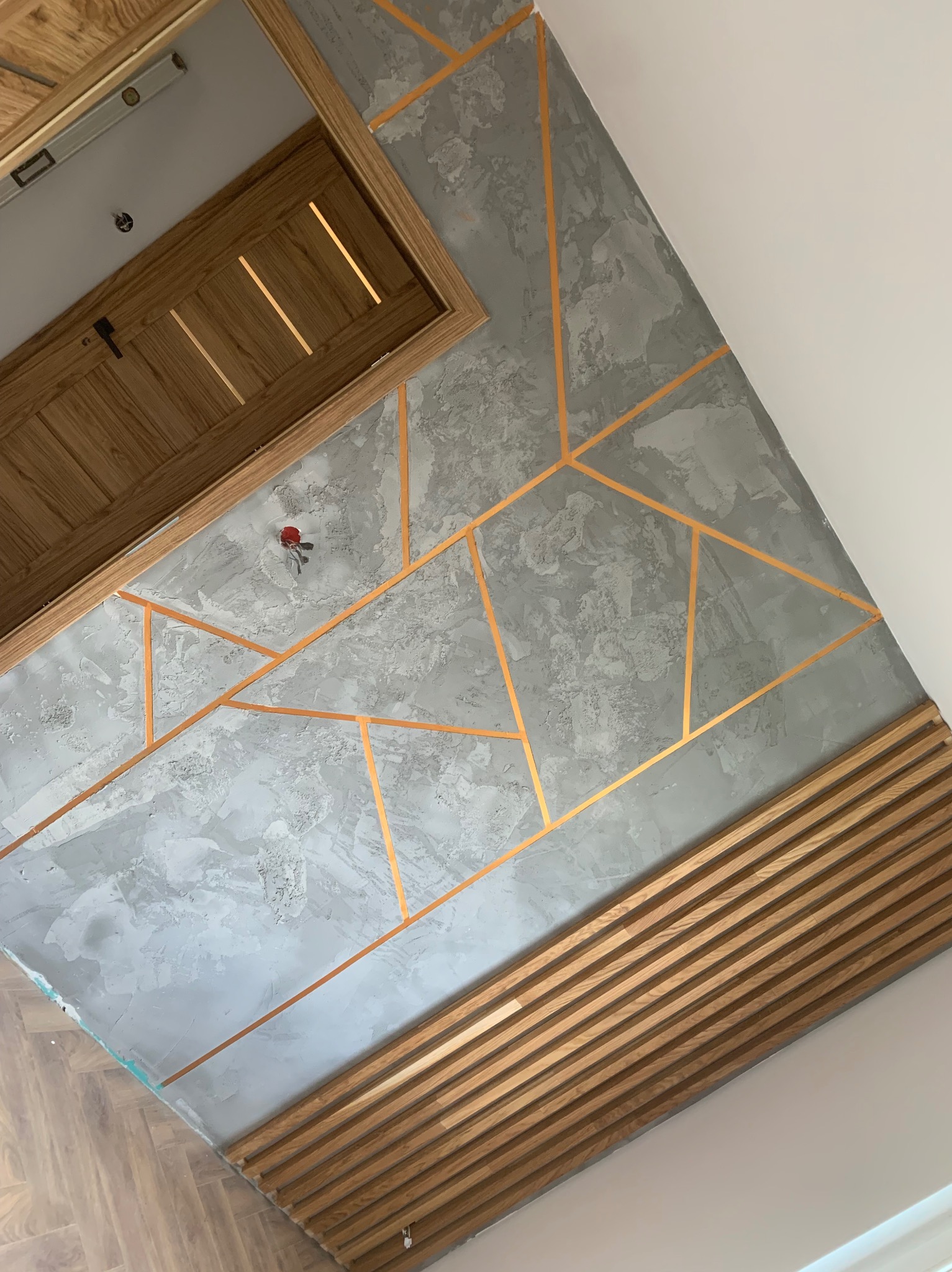 Dekoracyjna ściana z szarą fakturą, złotymi geometrycznymi liniami i drewnianymi panelami, widoczna także drewniana ościeżnica drzwiowa z poziomą listwą na górze.