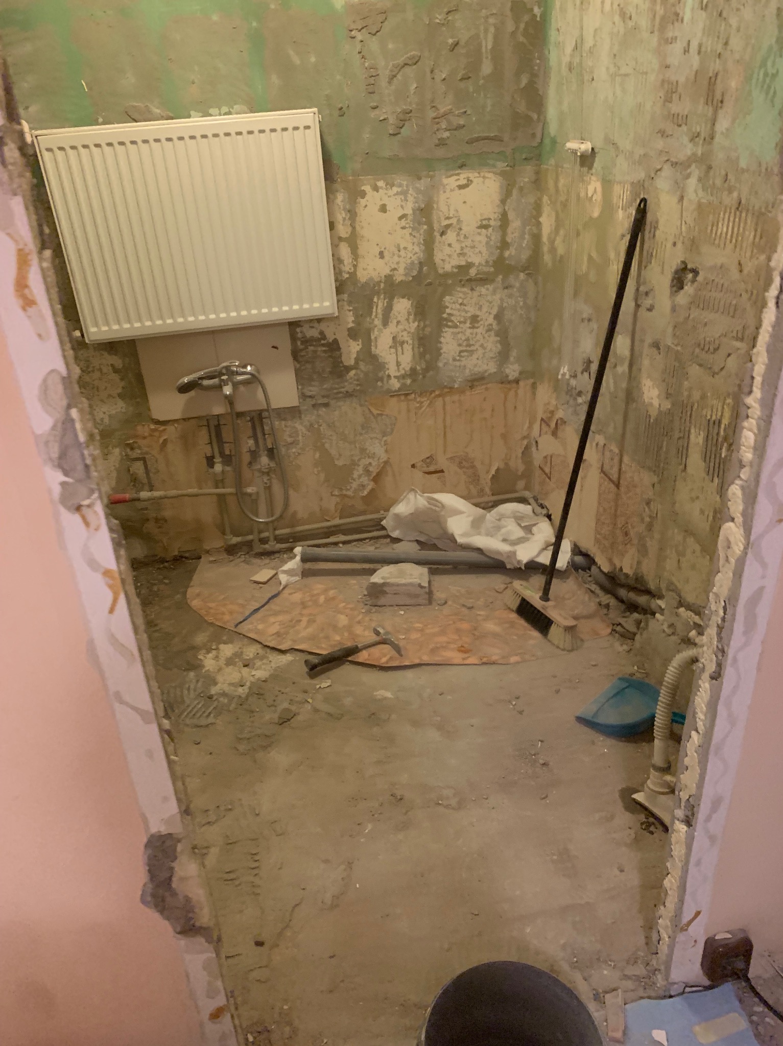 Łazienka w trakcie remontu: odsłonięte ściany, grzejnik na ścianie, rury, prowizoryczna podłoga, narzędzia i sprzęt budowlany, widoczne ślady po skuciu płytek.