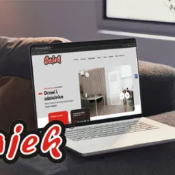 Laptop na szarej sofie wyświetla stronę internetową sklepu budowlanego Gajek z Lublina, prezentującą ofertę drzwi i ościeżnic.