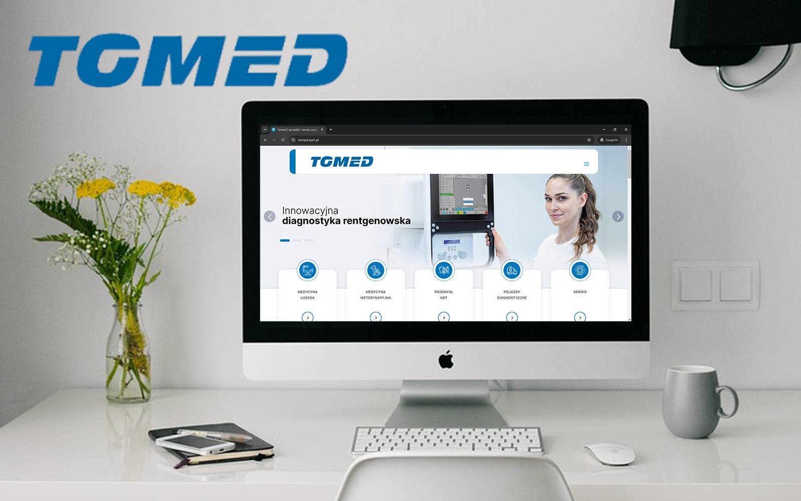 Ekran komputera iMac wyświetlający stronę internetową firmy TGMED na minimalistycznym białym biurku z bukietem kwiatów, notatnikiem i szarym kubkiem.