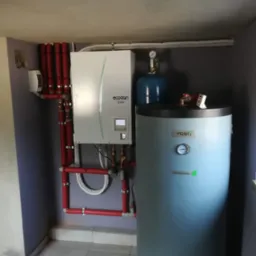 Pompa ciepła Ecodan Mitsubishi Electric z czerwonymi rurami izolowanymi i zbiornikiem Vasti w pomieszczeniu z szarą ścianą.