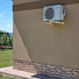 CHŁODER Kornel Gryboś - Zewnętrzna jednostka klimatyzacji Mitsubishi Electric zamontowana na elewacji domu z dekoracyjnym cokołem, widoczny fragment trawnika i huśtawka w tle.