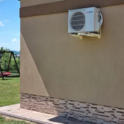 Zewnętrzna jednostka klimatyzacji Mitsubishi Electric zamontowana na elewacji domu z dekoracyjnym cokołem, widoczny fragment trawnika i huśtawka w tle.