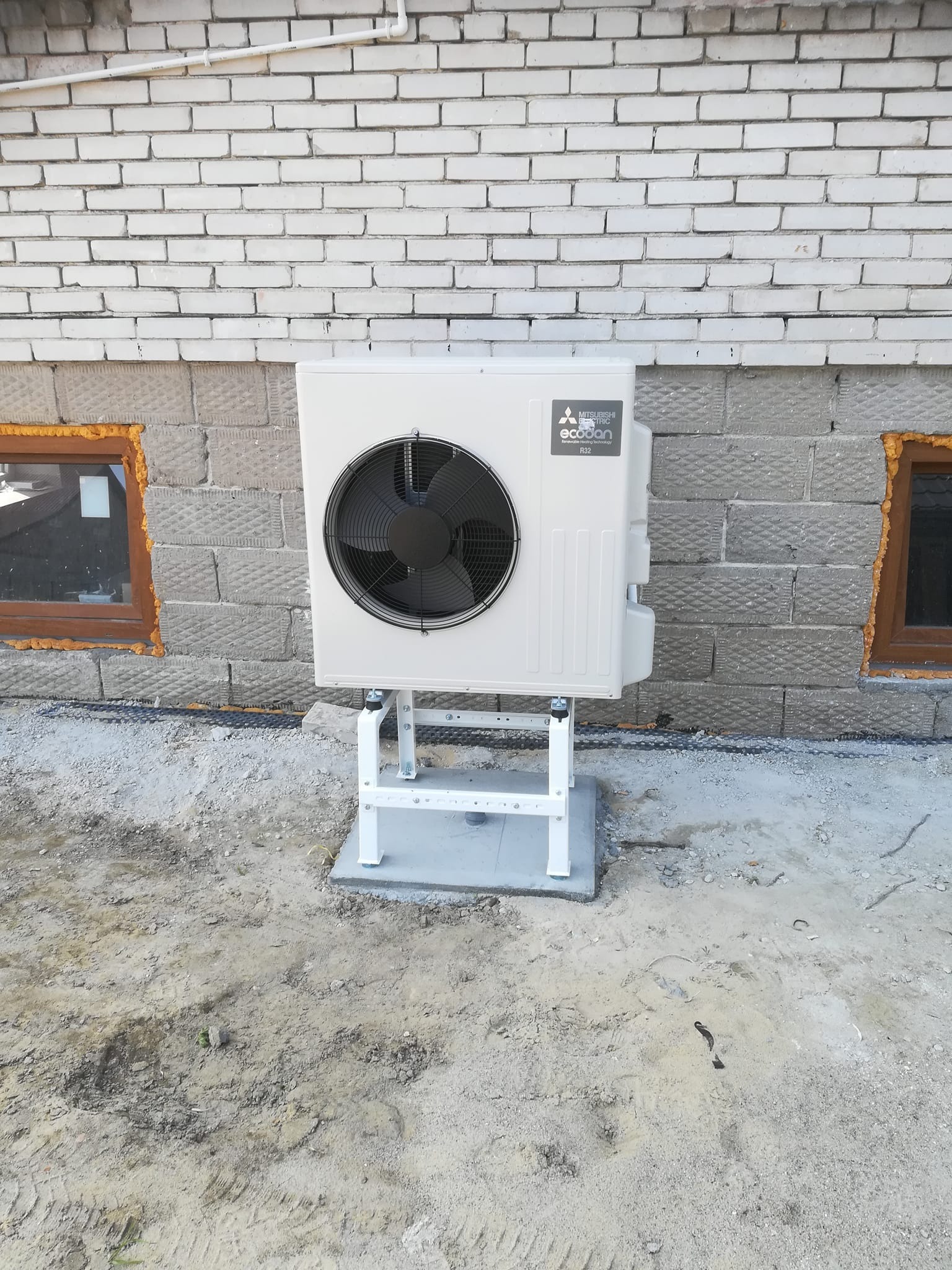Zewnętrzna jednostka pompy ciepła Mitsubishi Ecodan R32 zamontowana na białym stelażu na betonowej podstawie przy szarym, ceglanym murze budynku z oknami uszczelnionymi pianką montażową.