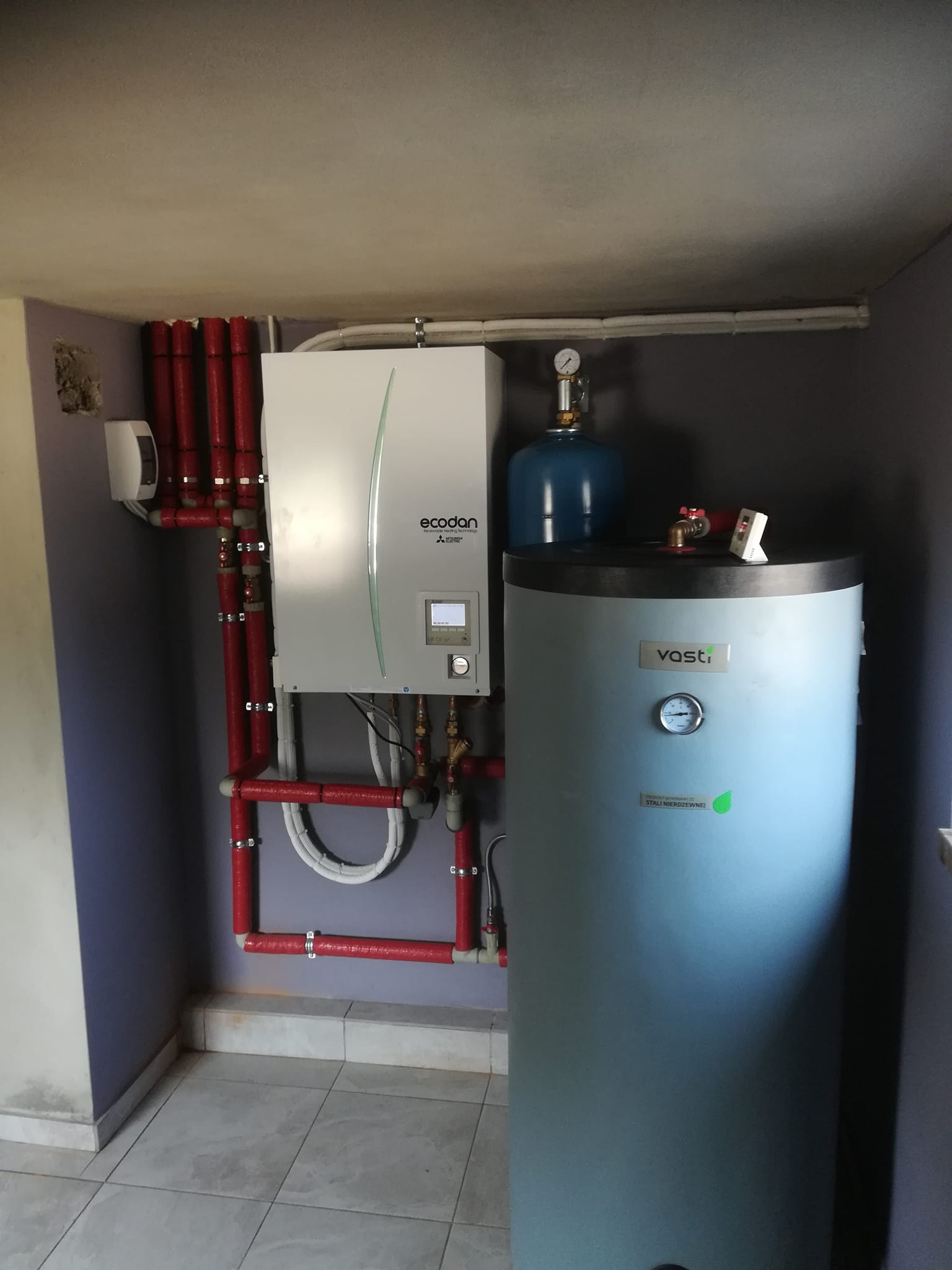 Pompa ciepła Ecodan Mitsubishi Electric z czerwonymi rurami izolowanymi i zbiornikiem Vasti w pomieszczeniu z szarą ścianą.
