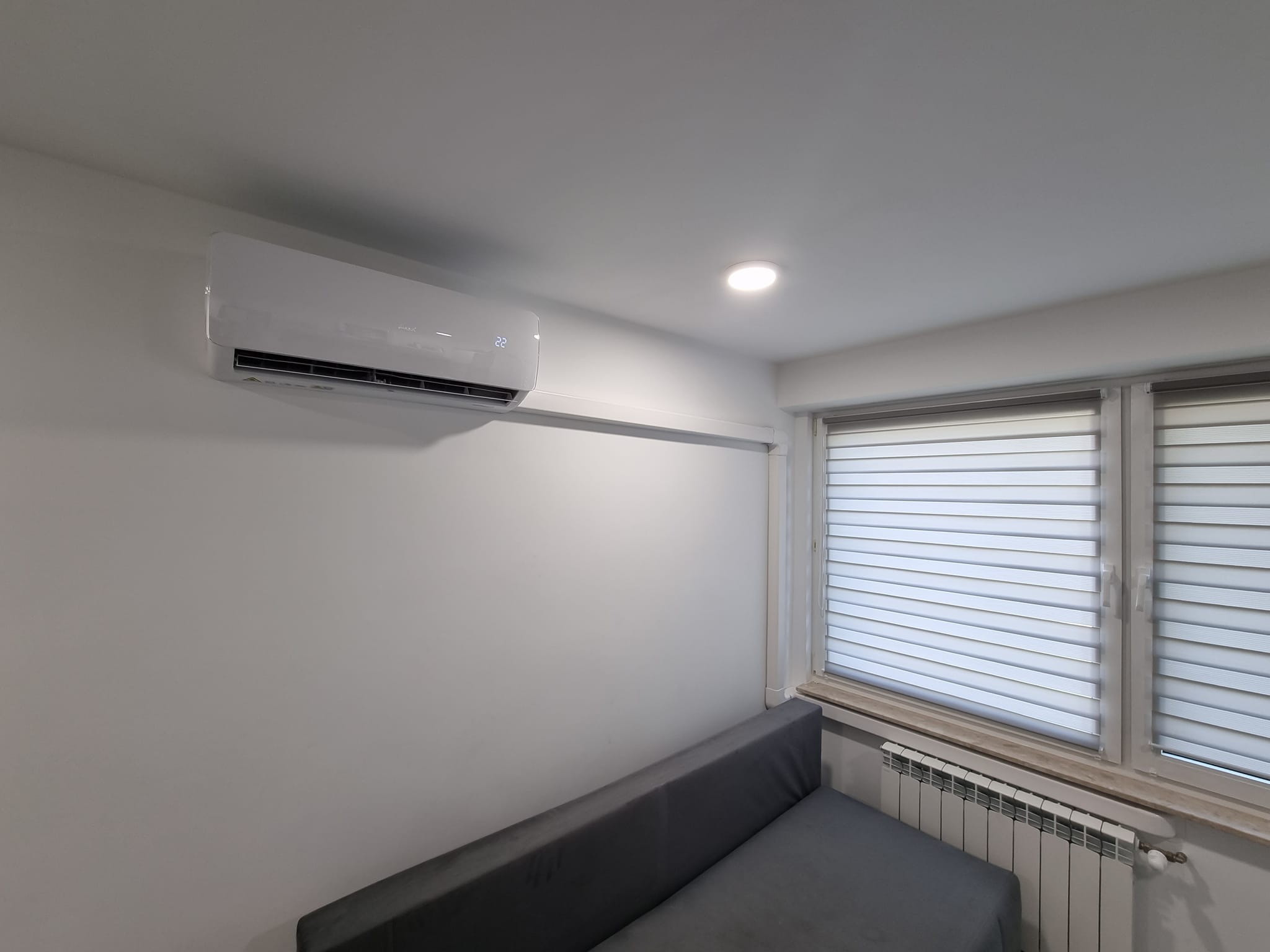 Biały klimatyzator ścienny w nowoczesnym salonie z szarym narożnikiem, widok z dołu, rolety okienne w tle.