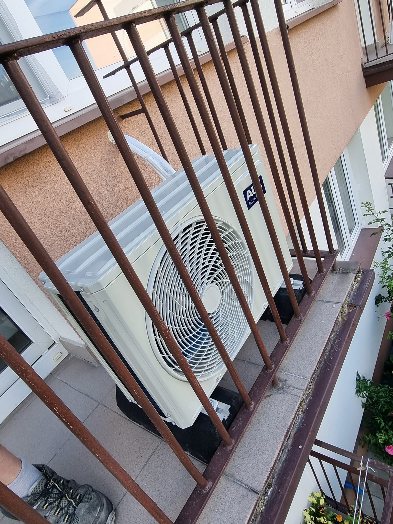 Biała jednostka zewnętrzna klimatyzacji zamontowana na balkonie, widoczna przez brązowe barierki, z fragmentem buta w kadrze.