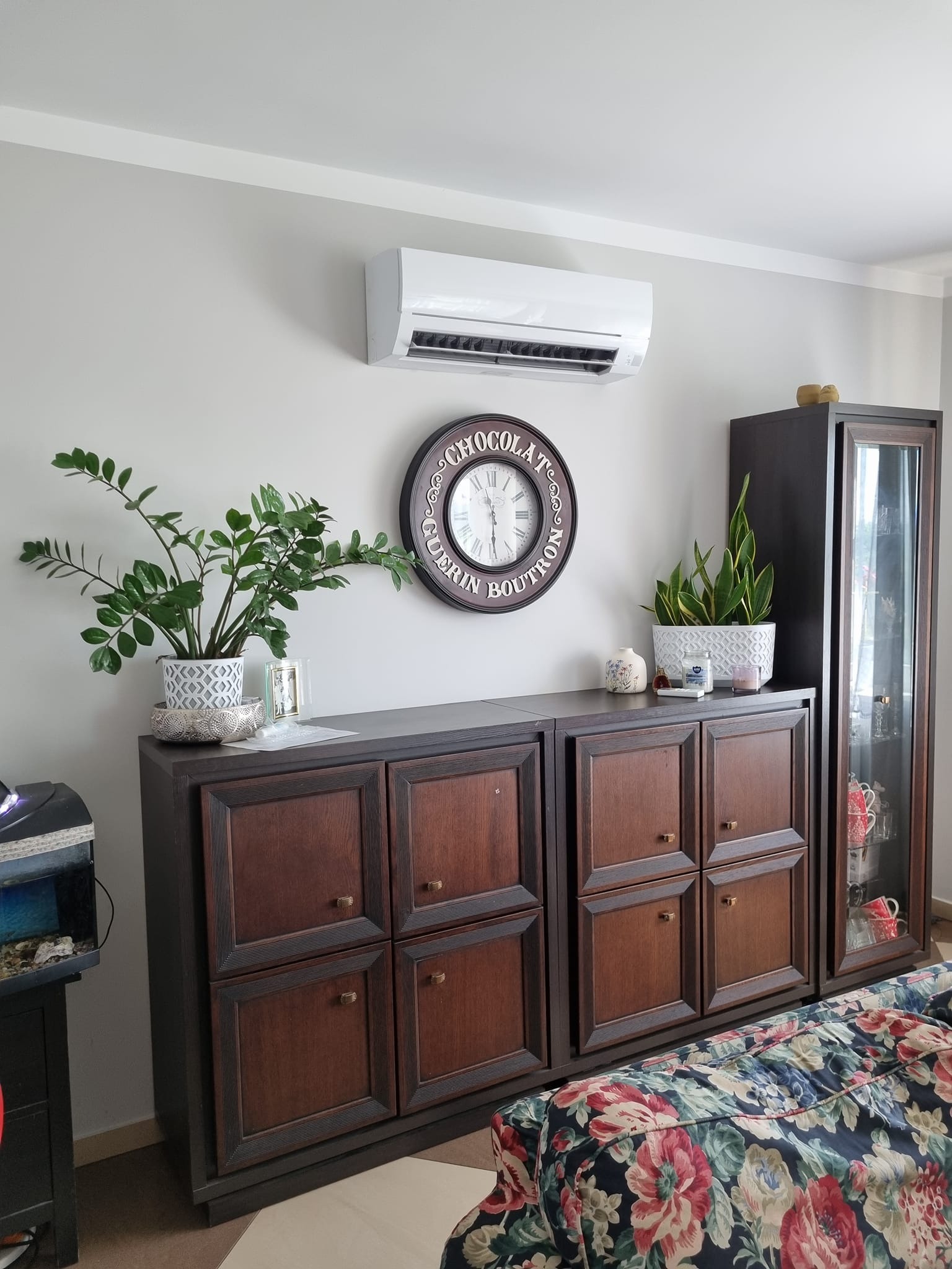 Biała jednostka klimatyzacyjna zamontowana na ścianie nad ciemnobrązowymi szafkami, ozdobiona roślinami doniczkowymi i zegarem ściennym.