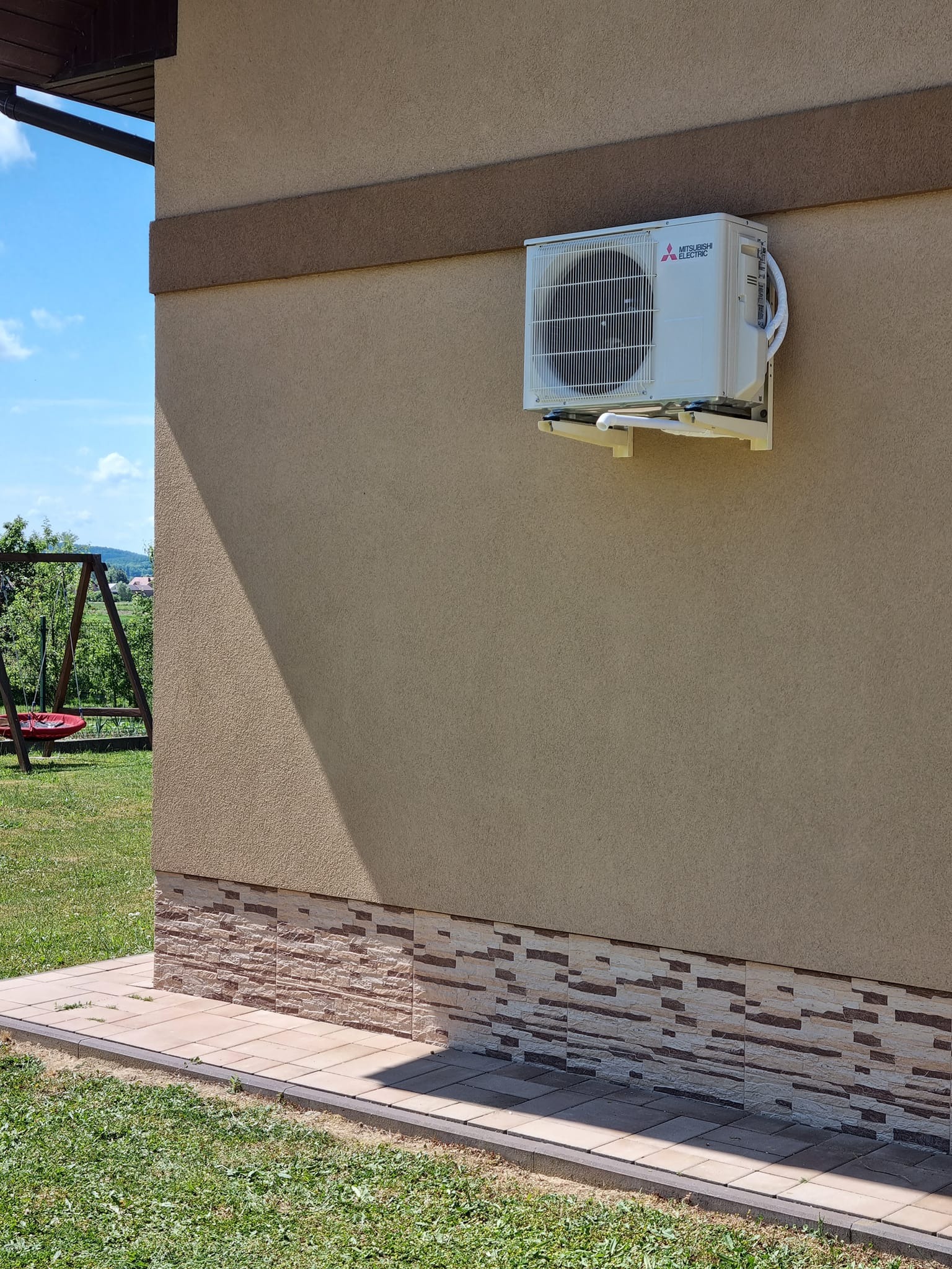Zewnętrzna jednostka klimatyzacji Mitsubishi Electric zamontowana na elewacji domu z dekoracyjnym cokołem, widoczny fragment trawnika i huśtawka w tle.