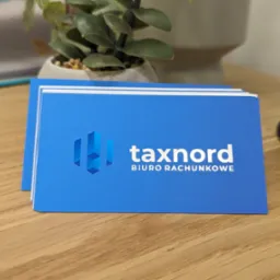 Stos niebieskich wizytówek z białym logo i napisem 'taxnord BIURO RACHUNKOWE' leży na drewnianym biurku, obok doniczki z rośliną i czarnego kosza na śmieci.