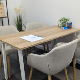 Jasny pokój biurowy z drewnianym stołem otoczonym czterema krzesłami, na stole leży biały dokument i niebieski długopis, w tle regał z drewnianymi szufladkami i roślina w doniczce.