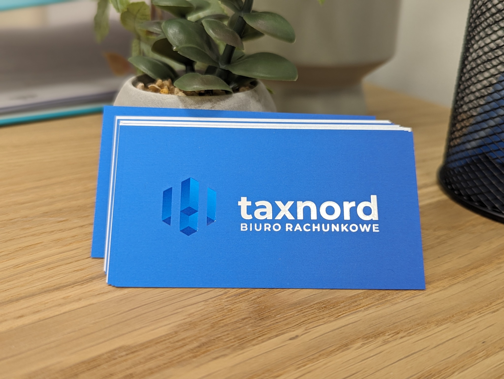 Stos niebieskich wizytówek z białym logo i napisem 'taxnord BIURO RACHUNKOWE' leży na drewnianym biurku, obok doniczki z rośliną i czarnego kosza na śmieci.