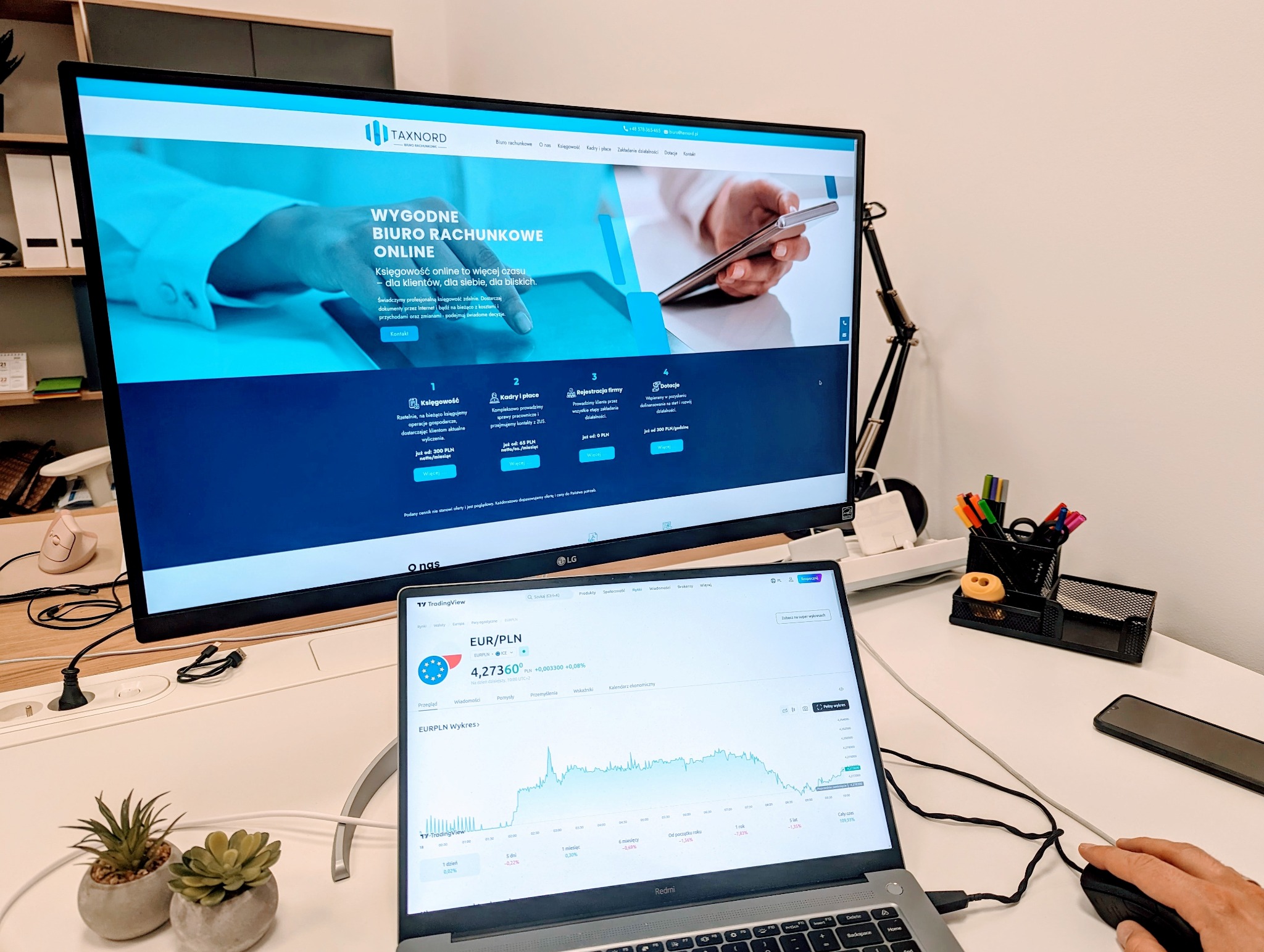 Monitor wyświetla stronę internetową biura rachunkowego Taxnord z ofertą księgowości online, obok laptop z wykresem kursu EUR/PLN, na biurku doniczki z sukulentami, organizer na długopisy i myszka...