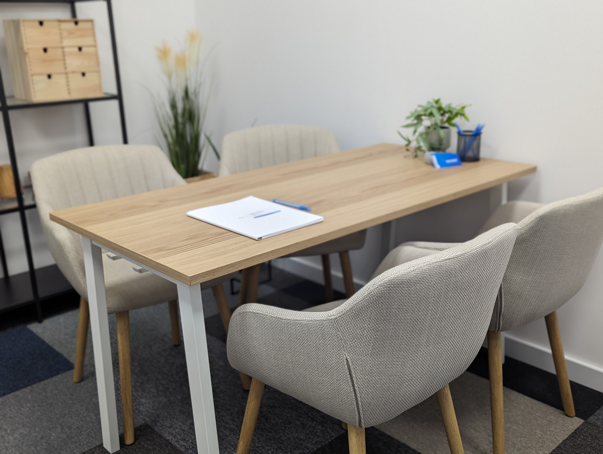 Jasny pokój biurowy z drewnianym stołem otoczonym czterema krzesłami, na stole leży biały dokument i niebieski długopis, w tle regał z drewnianymi szufladkami i roślina w doniczce.