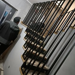 TRÓJSPAW - Wnętrze klatki schodowej w trakcie montażu metalowej konstrukcji schodów z czarnymi, pionowymi balustradami, widoczny pracownik w kurtce i czapce oraz częściowo widoczny drugi pracownik na schodach.
