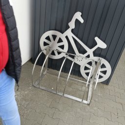 TRÓJSPAW - Metalowy stojak na rowery z dekoracyjnym motywem roweru, umieszczony na szarym bruku przed ciemnoszarą ścianą.