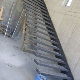 TRÓJSPAW - Surowe, metalowe schody w trakcie montażu w surowym wnętrzu, obok stoi niebieska puszka z farbą.