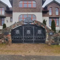 Kuta, dwuskrzydłowa brama wjazdowa z motywem lwa, wkomponowana w kamienny mur, z numerem domu '10', na tle dwupiętrowego domu z cegły i białego tynku.