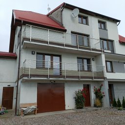 TRÓJSPAW - Trzykondygnacyjny dom z balkonami z balustradami ze stali nierdzewnej, czerwonym dachem, brązowymi oknami i drzwiami garażowymi, oraz krzewami róż pnących się po ścianie.