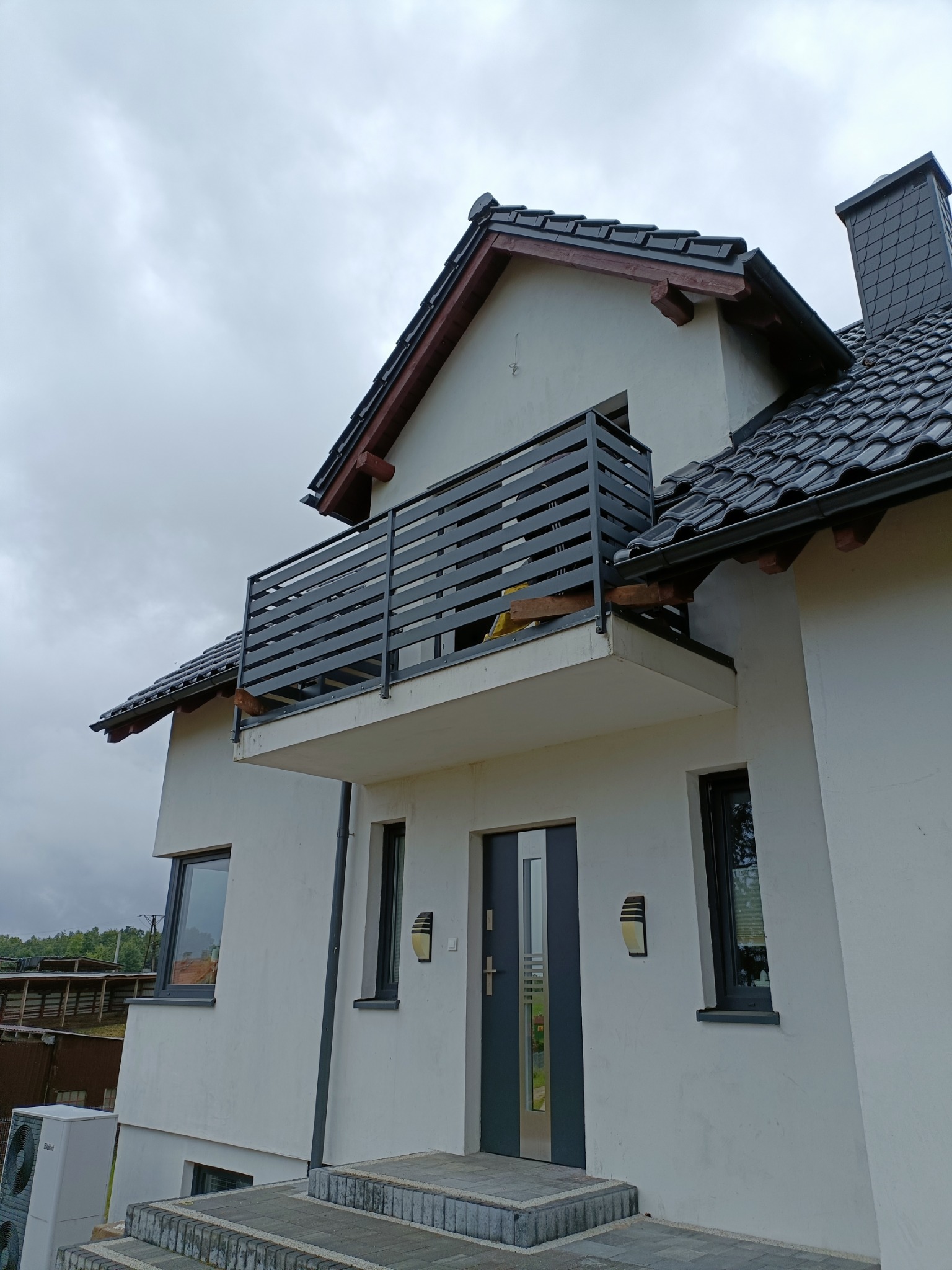 Nowoczesny dom z balkonem zabezpieczonym czarną, poziomą balustradą metalową, widok z dołu na szare płytki dachowe i białą elewację w pochmurny dzień.