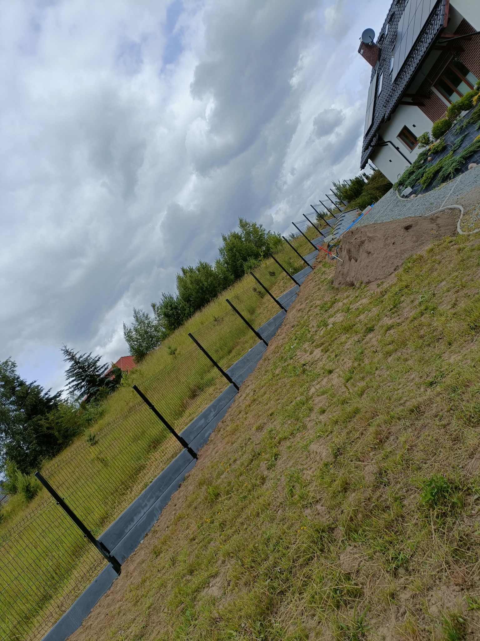 Montaż ogrodzenia panelowego na nierównym terenie, widoczne słupki i siatka w trakcie instalacji, w tle dom i pochmurne niebo.