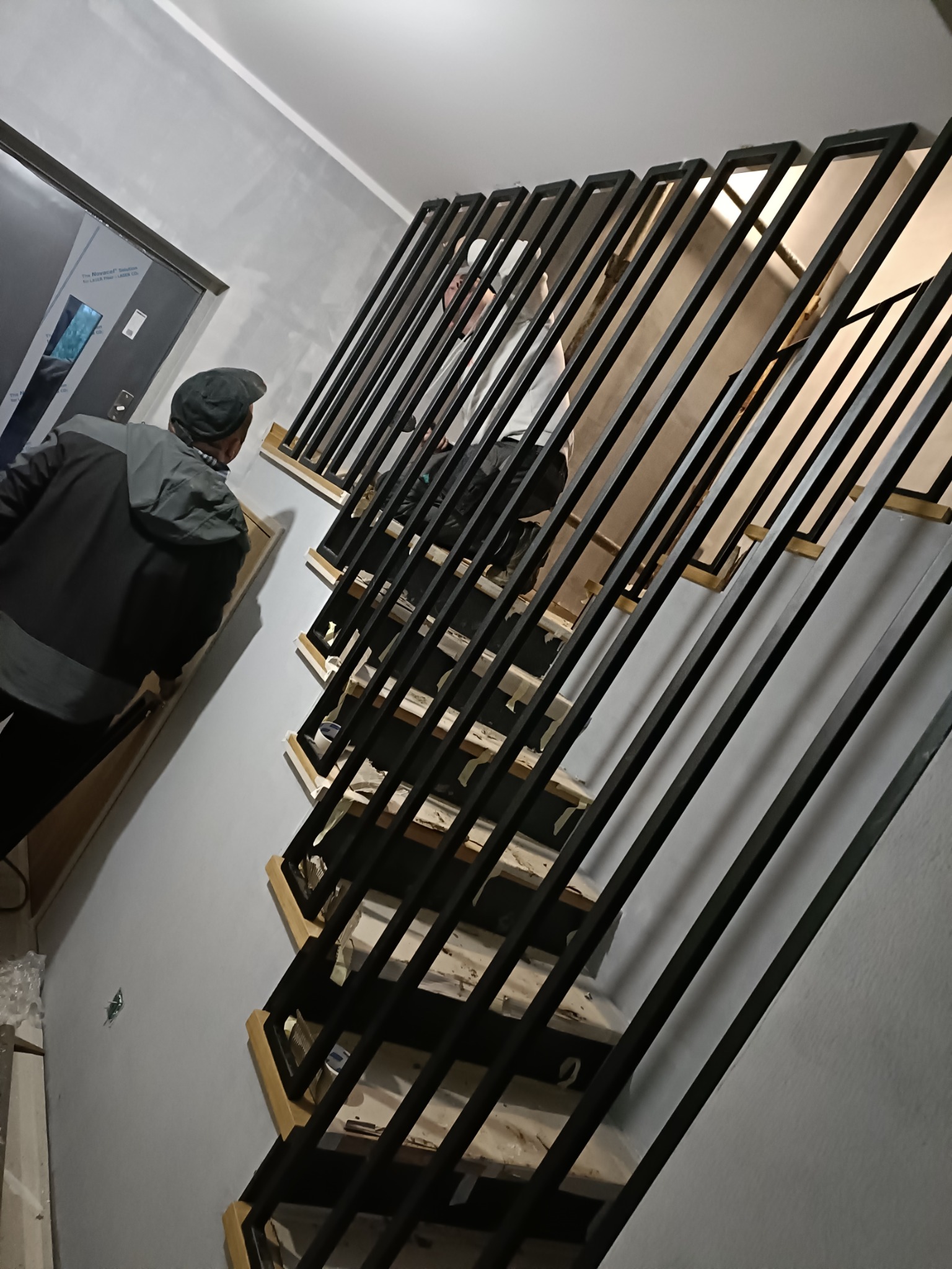 Wnętrze klatki schodowej w trakcie montażu metalowej konstrukcji schodów z czarnymi, pionowymi balustradami, widoczny pracownik w kurtce i czapce oraz częściowo widoczny drugi pracownik na schodach.