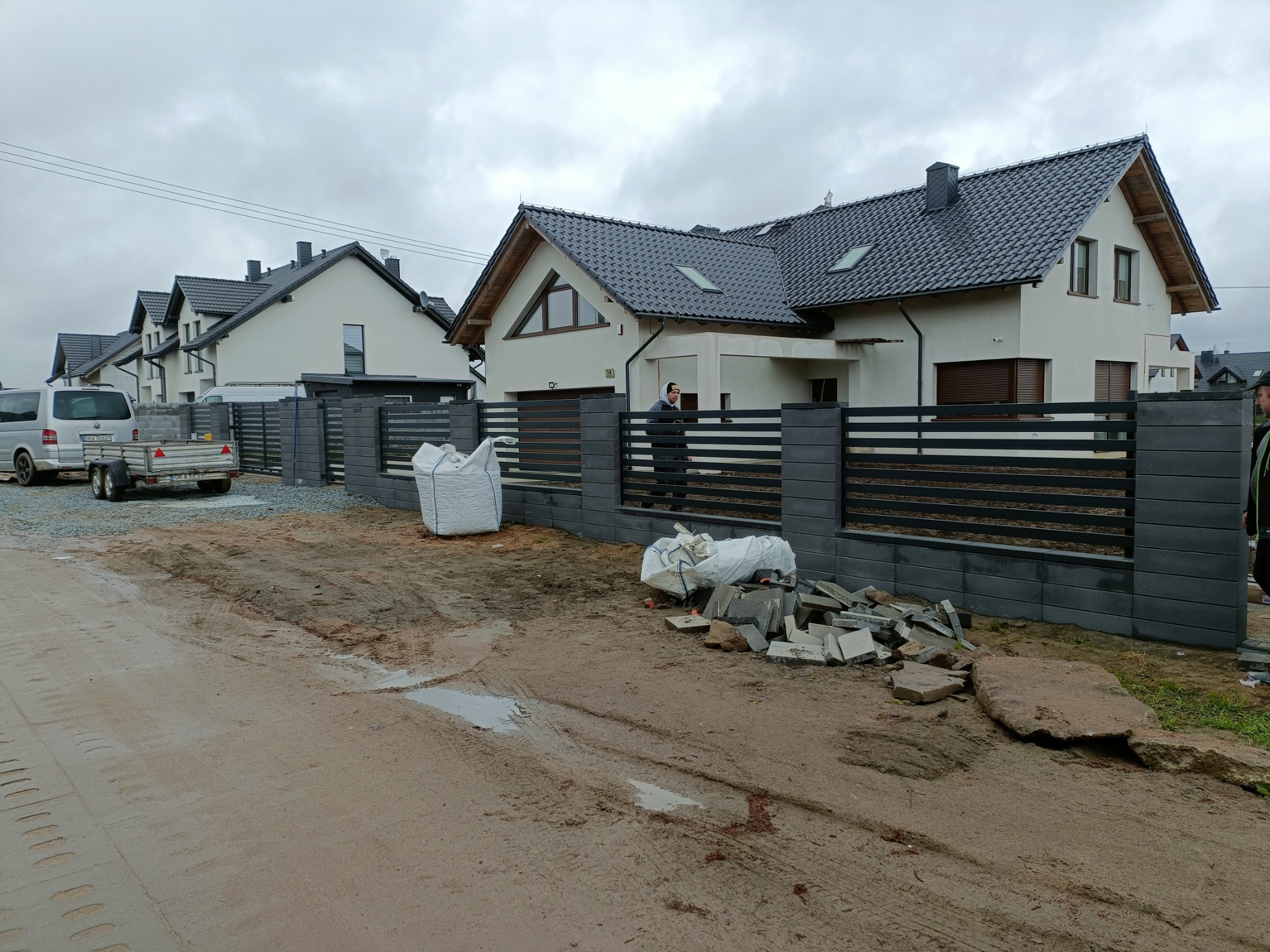 Nowoczesne ogrodzenie panelowe z grafitowych elementów betonowych i poziomych przęseł, widoczne w trakcie montażu przy nowo wybudowanym osiedlu domów jednorodzinnych, z materiałami budowlanymi...